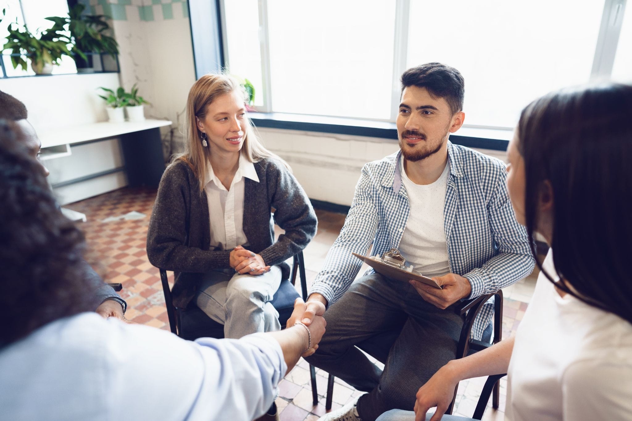 A group therap session with a man sitting on a chair and holding some document shaking hands with a woman